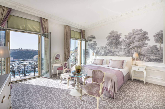A luxurious bedroom features a neatly made bed, elegant chairs, and a glass table. Large windows open to a scenic view, while a mural depicting trees adorns the wall.