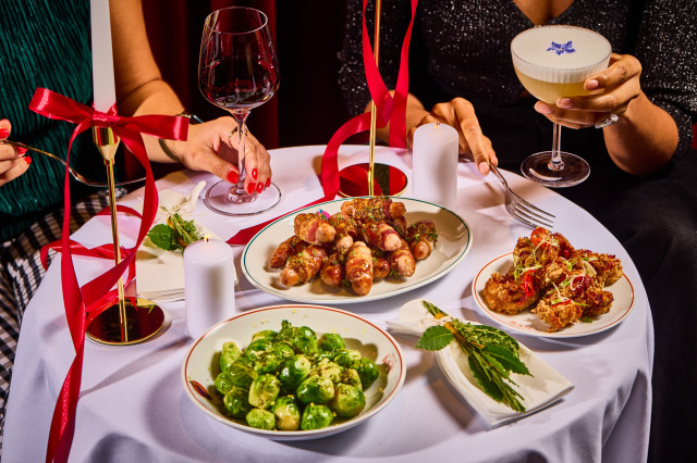 A small table displays festive appetizers including Brussels sprouts, bacon-wrapped sausages, and fried bites. Two people toast with wine and a cocktail amid candles and ribbon decor.