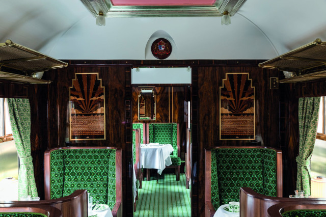 Luxurious train carriage, featuring art deco green seats and ornate wood paneling, is set for dining with white tablecloths, under soft natural light from curtained windows.