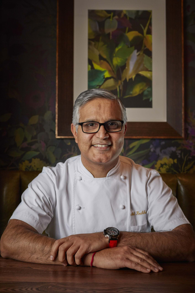 A chef, wearing a white coat and glasses, sits smiling at a wooden table