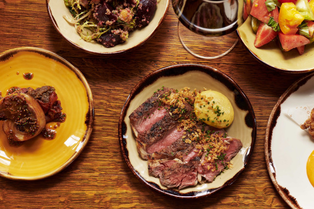 A wooden table holds assorted dishes: sliced steak with garnish, hearty stew, fresh vegetable salad, quinoa mix, and a glass of red wine, creating a rich dining scene