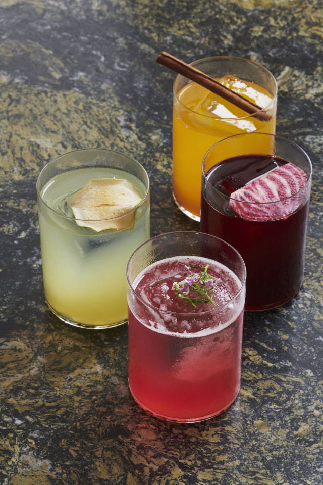 Four colorful cocktails are arranged on a marbled surface. Each drink is in a glass, garnished distinctively with items like cinnamon, herbs, or fruit slices, adding a decorative touch.