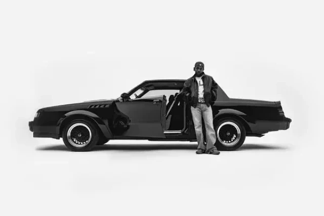 A classic black car with an open driver's door stands against a white background. A man in a leather jacket and jeans leans casually on it.