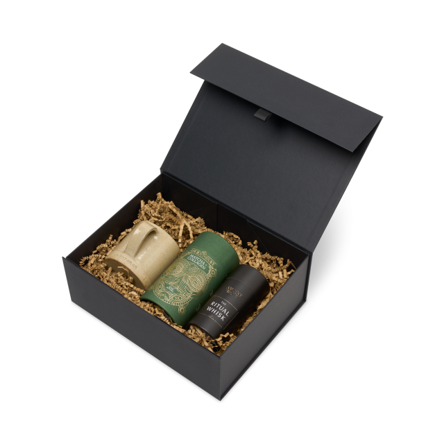 A black gift box contains three items on bed of shredded paper: a beige ceramic mug, a green package, and a black container labeled