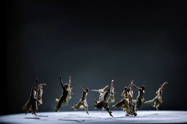 Dancers in flowing costumes leap energetically on a dimly lit stage, creating a sense of dynamic movement and rhythm within a spotlighted performance space.