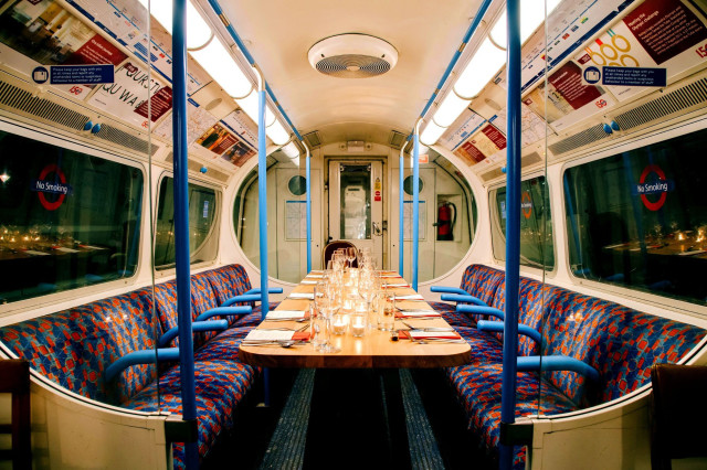 A wooden dining table set with glasses and cutlery is placed inside a retro train carriage with patterned seats, lit warmly. Surrounding advertisements and 