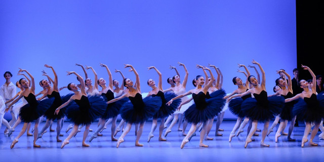 Ballet dancers perform synchronized movements on stage. The dancers wear blue tutus and strike elegant poses with raised arms in a well-lit theater setting, creating a visually harmonious scene.