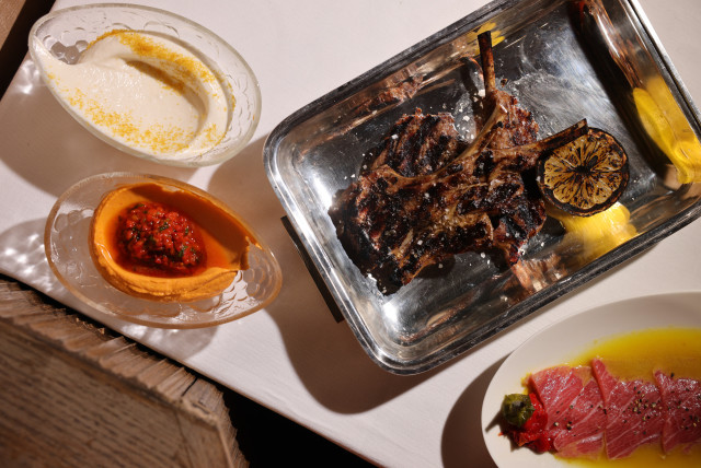 A metal tray holds grilled lamb chops with a charred lemon slice. Surrounding it are dishes of hummus, red pepper dip, and thinly sliced fish on a white tablecloth.