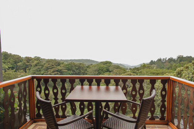 A wooden table with two wicker chairs sits on a balcony overlooking a lush, green forested landscape under a clear sky.