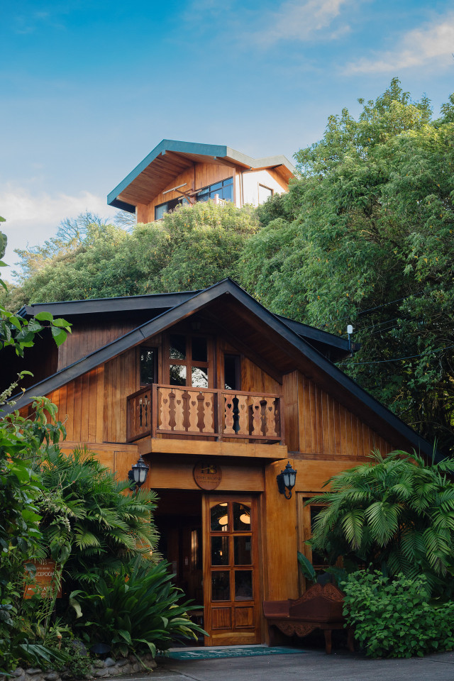 A wooden house with a small balcony stands among lush greenery under a clear blue sky. A sign reads 