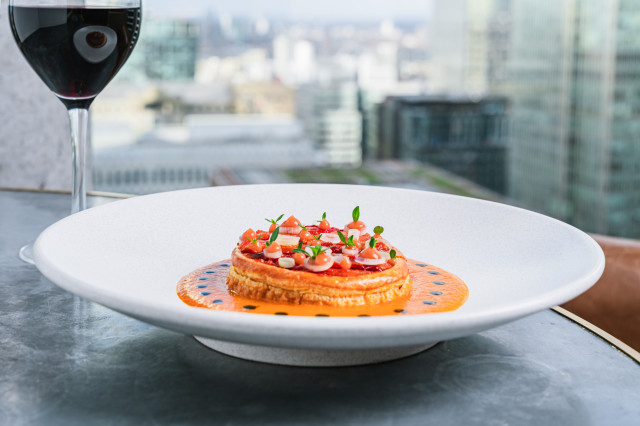 A gourmet dish is elegantly plated with vibrant toppings on a white plate, placed on a table. A glass of red wine sits nearby, overlooking a cityscape.
