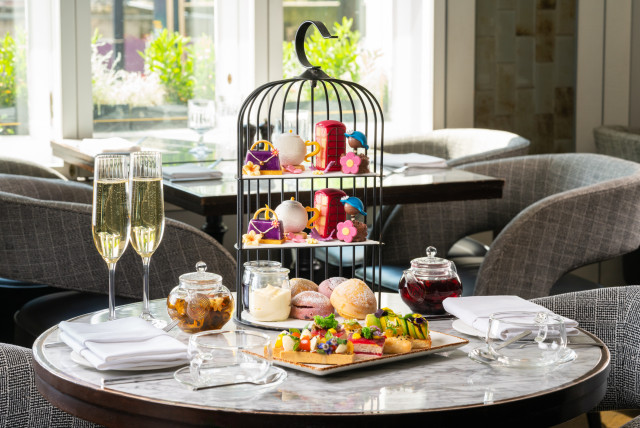 A birdcage displays colorful pastries, surrounded by a table set with champagne glasses and assorted desserts in glass jars, within a bright, elegant dining area with modern chairs.