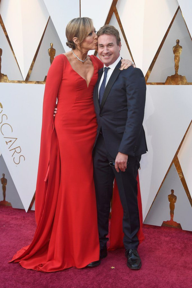 A woman in a red gown kisses a smiling man in a suit on the cheek