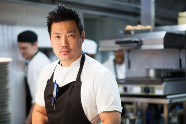 Chef stands in a restaurant kitchen, wearing a white shirt and black apron with a pen clipped on