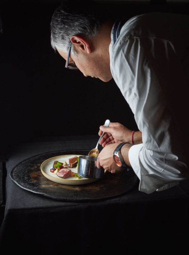 A chef meticulously garnishes a plated dish with sauce from a small pot, under soft lighting, focused on precision and presentation in an elegant, dark culinary setting