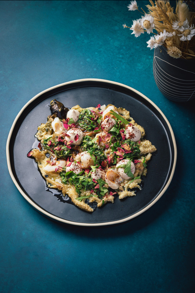 A black plate holds a colorful dish with roasted vegetables, creamy sauce, and fresh herbs. The setting is a teal surface beside a black vase with dried flowers.