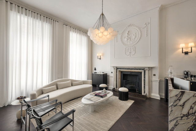 Sculpted ceiling chandelier hangs above a modern living room with a curved sofa, oval coffee table, and fireplace. Tall draped windows and minimalist decor enhance the elegant atmosphere.