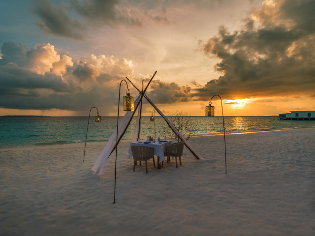 A beachfront dining setup with a table and chairs under a canopy, surrounded by lanterns, is set against a vibrant ocean sunset and scattered clouds.