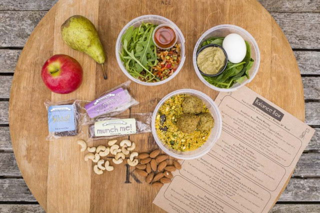 A wooden table displays a variety of foods: a pear, an apple, mixed salad with dressing, a boiled egg with greens and dip, falafel with couscous, snack bars, nuts, and a 