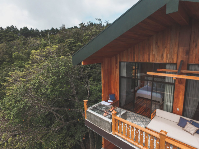 A wooden house with large windows extends over a lush forest. A person relaxes on a balcony daybed under cloudy skies, providing a serene, secluded atmosphere.