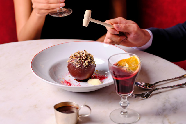 A chocolate dessert sphere sits on a white plate, being tapped with a small hammer. Nearby are a glass containing a drink and a small metal cup, all on a marble table surface.