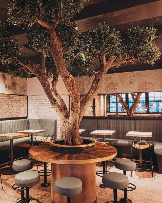 A large tree grows through the center of a round table, surrounded by stools in a cozy restaurant setting with brick walls and windows
