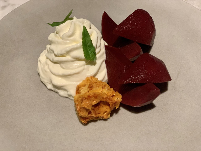 A dessert plate features whipped cream with green leaf garnish, alongside sliced red fruit and a piece of honeycomb, set on a neutral-colored plate