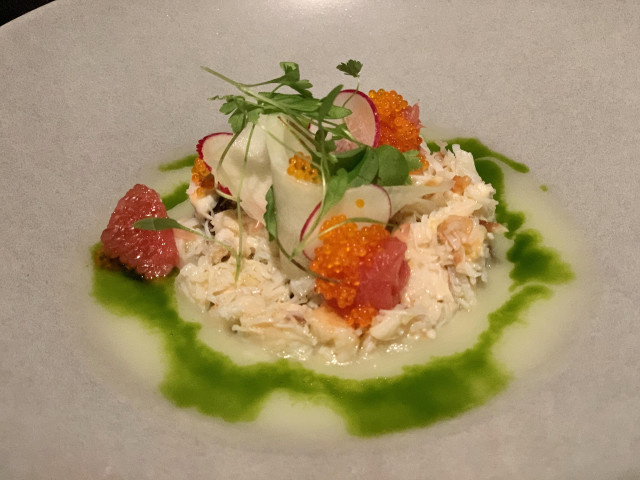 Crab salad garnished with grapefruit segments, radish slices, fish roe, and microgreens sits on a white plate, surrounded by a vibrant green sauce, in a fine dining setting