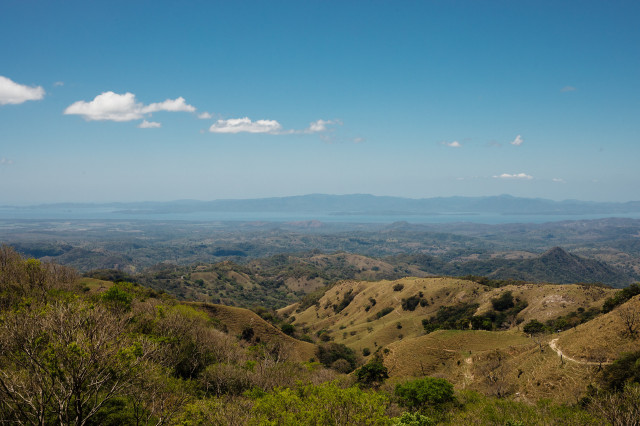 Rolling hills stretch across a landscape dotted with sparse vegetation, leading to a distant, calm body of water under a clear blue sky with a few scattered clouds.