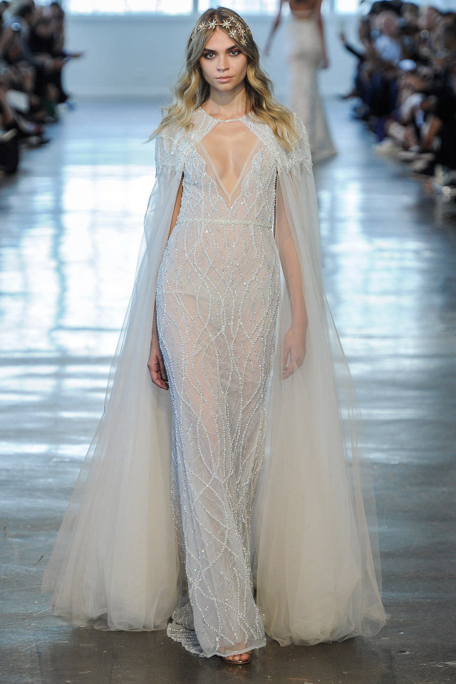 A model walks in a sheer, embellished gown with a flowing cape, under soft lighting, on a runway lined with seated spectators