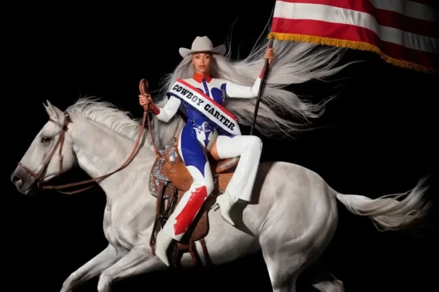 A person in a cowboy outfit rides a white horse, holding a flag. They wear a hat and a 