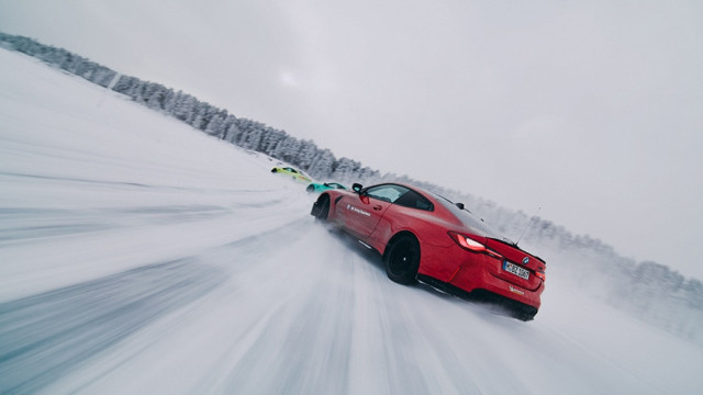 A red sports car drifts on a snow-covered track, closely followed by two others. Snow swirls around, and trees line the horizon. Text on the car reads 