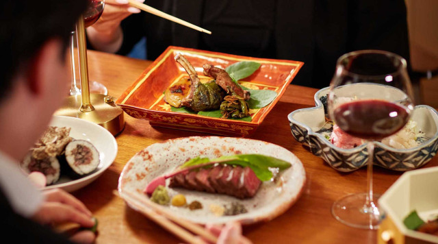 A table is set with elegantly plated dishes, including sushi, steak, and vegetables, accompanied by two glasses of red wine. People use chopsticks, creating a fine dining atmosphere.