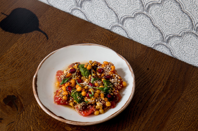 A white bowl contains a colorful salad with grains, vegetables, and greens, placed on a wooden table. The backdrop features patterned wallpaper, creating a warm, inviting setting.