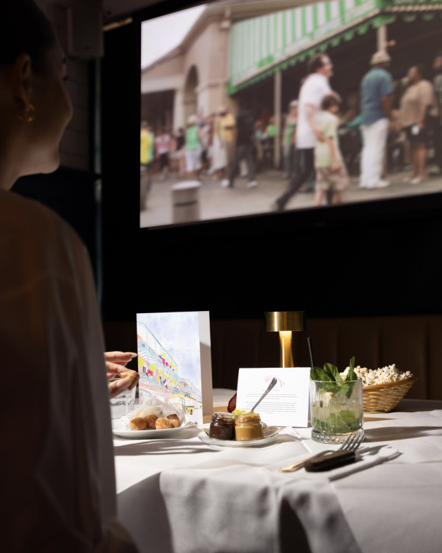 A person sits at a white-draped table adorned with desserts, a mint drink, and a lamp, watching a crowded scene on a large screen in a dimly lit setting.