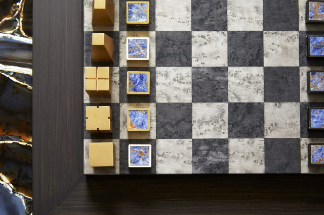 Chess pieces with abstract geometric designs are arranged on a checkered board, positioned on a wooden table with a decorative, marbled edge
