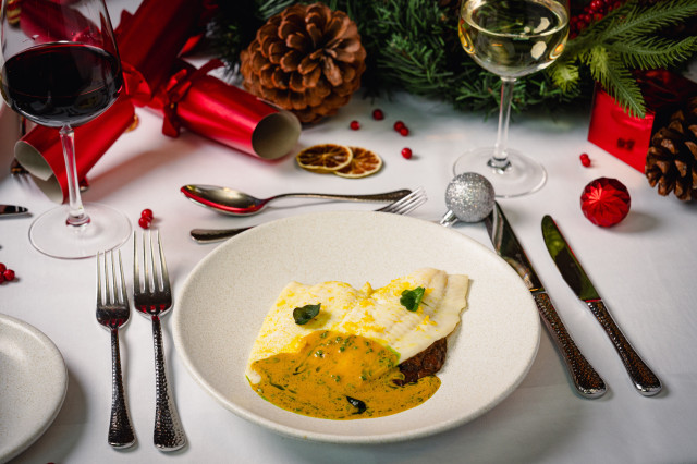 A dish of fish with sauce is on a festive dining table. Nearby are wine glasses, cutlery, a red cracker, pinecones, and evergreen branches, creating a holiday atmosphere.