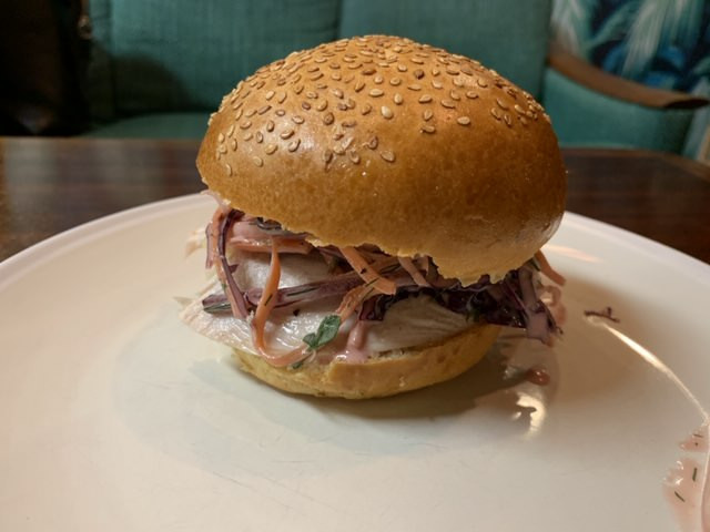 A sesame seed bun holds a sandwich filled with sliced meat, shredded vegetables, and dressing, resting on a white plate