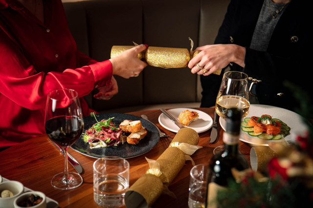 Two people pull a gold Christmas cracker across a dining table, set with wine glasses, salads, and holiday decorations, creating a festive atmosphere.