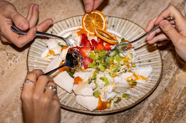 A dessert plate with whipped cream, strawberries, kiwi, orange slices, and sauce is being shared by three people using spoons, atop a textured stone surface.