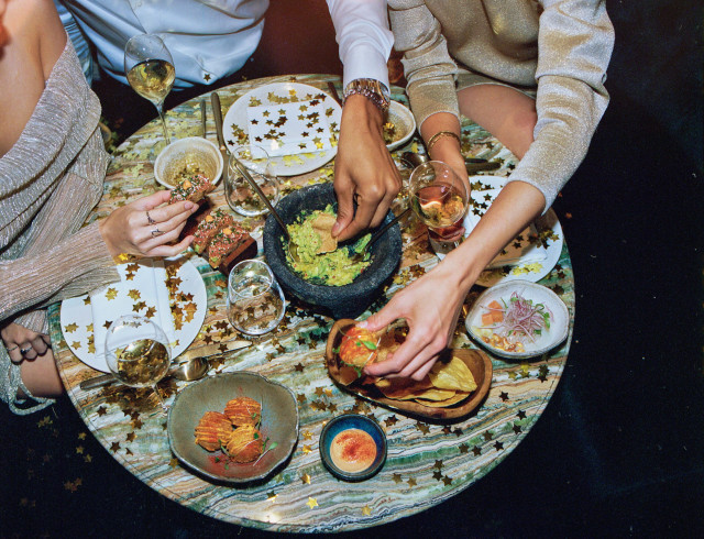 Guacamole is being shared at a festive table, surrounded by plates with food, drinks, and gold star confetti. Three people are reaching for food and drinks, creating a celebratory atmosphere.