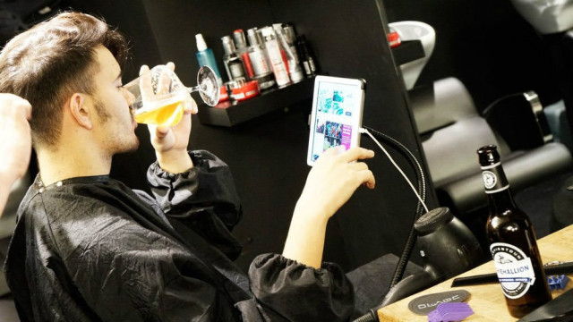 A person in a barber's chair drinks beer while viewing a tablet