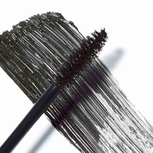 A black mascara brush smears dark mascara across a white surface, showcasing the bristles and the texture of the applied product