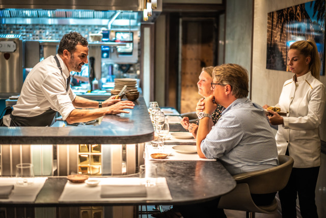 Chef interacts with diners seated at a stylish bar counter in a modern, warmly-lit restaurant. A server in white holds a tray, attending to the guests.