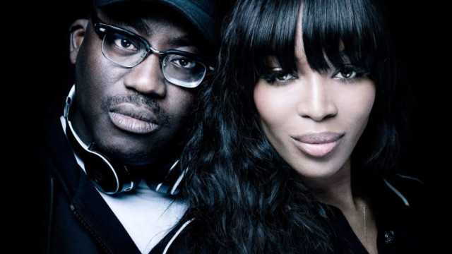 Two individuals, one wearing glasses and a cap with headphones around the neck and the other with long, dark hair, are posed closely together against a dark background