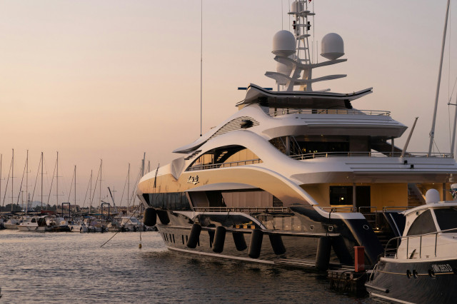 A large luxury yacht is docked at a marina during sunset, surrounded by smaller boats and masts, with calm water reflecting the evening light.