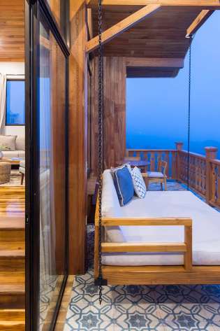 Wooden swing bed hangs on a chain, overlooking a misty mountain view. It's on a tiled balcony attached to a cozy room with wooden floors and warm lighting.
