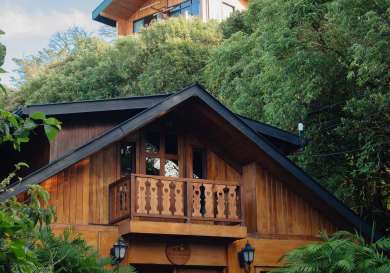 A wooden house with a small balcony stands among lush greenery under a clear blue sky. A sign reads 