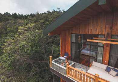 A wooden house with large windows extends over a lush forest. A person relaxes on a balcony daybed under cloudy skies, providing a serene, secluded atmosphere.
