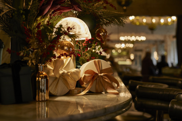 Gift-wrapped boxes with ribbons sit on a marble bar, surrounded by festive greenery and warm lighting, creating a cozy, holiday atmosphere in a dimly lit restaurant interior.